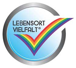 Eine rundes, silbeners Emblem, darin ein blauer Verlauf und ein großer Haken in Regenbogenfarben, darüber geschrieben steht: Lebensort Vielfalt®